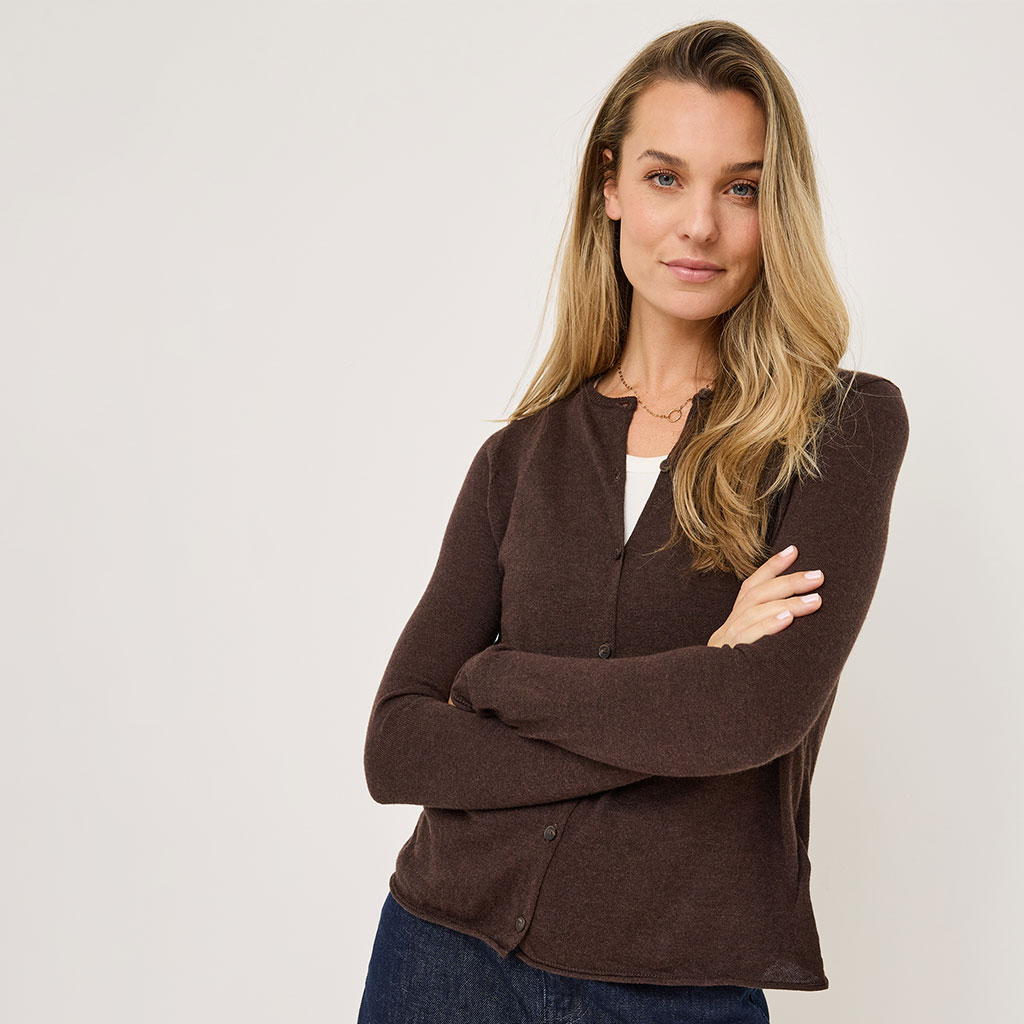 Ritratto di una persona dai lunghi capelli biondi con un cardigan marrone su uno sfondo chiaro, con le braccia incrociate.