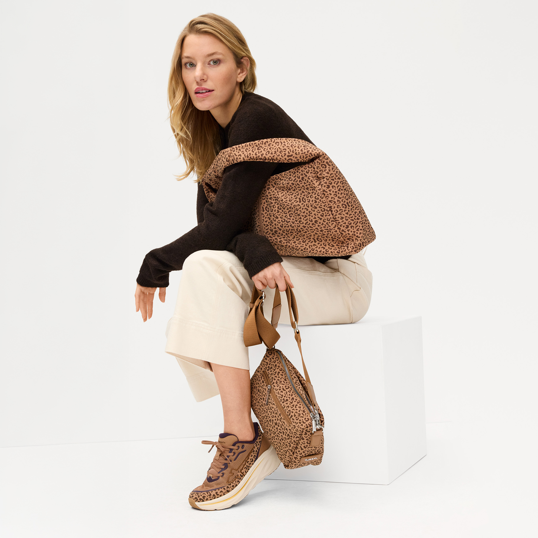 Vrouw zit op een kubus, draagt een donkere trui, cr&egrave;mekleurige broek, sneakers met luipaardprint en houdt een tas met luipaardmotief vast.