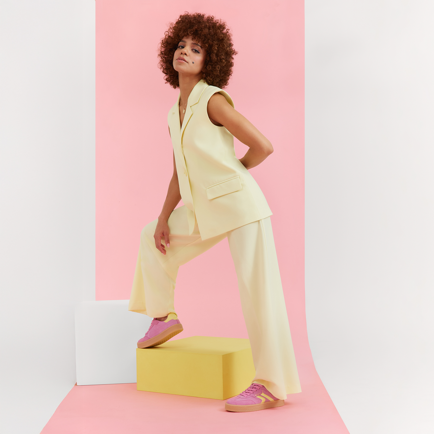 Model in cream suit and pink sneakers poses dynamically on geometric pedestals against pink background