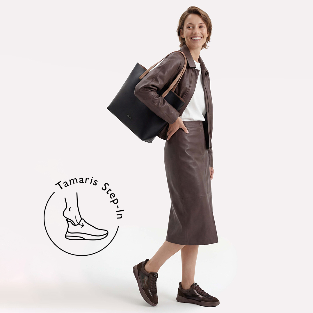Glimlachende vrouw in een bruine leren jas en rok met zwarte tas en bruine sneakers. Logo: Tamaris Step-In.