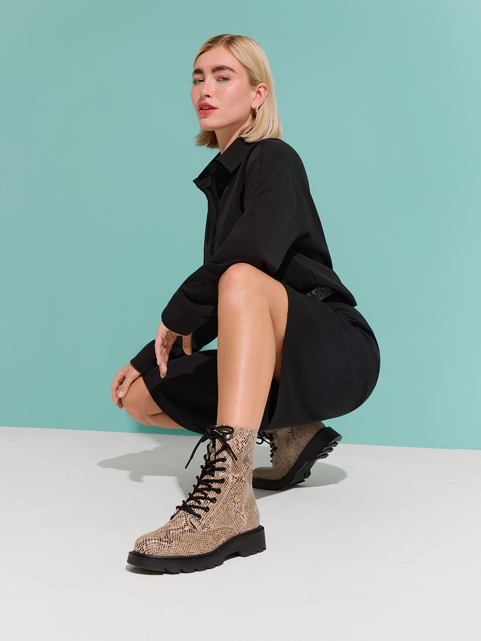 Modella in abito nero e stivaletti stringati in pelle di serpente beige posa accovacciata davanti a uno sfondo turchese