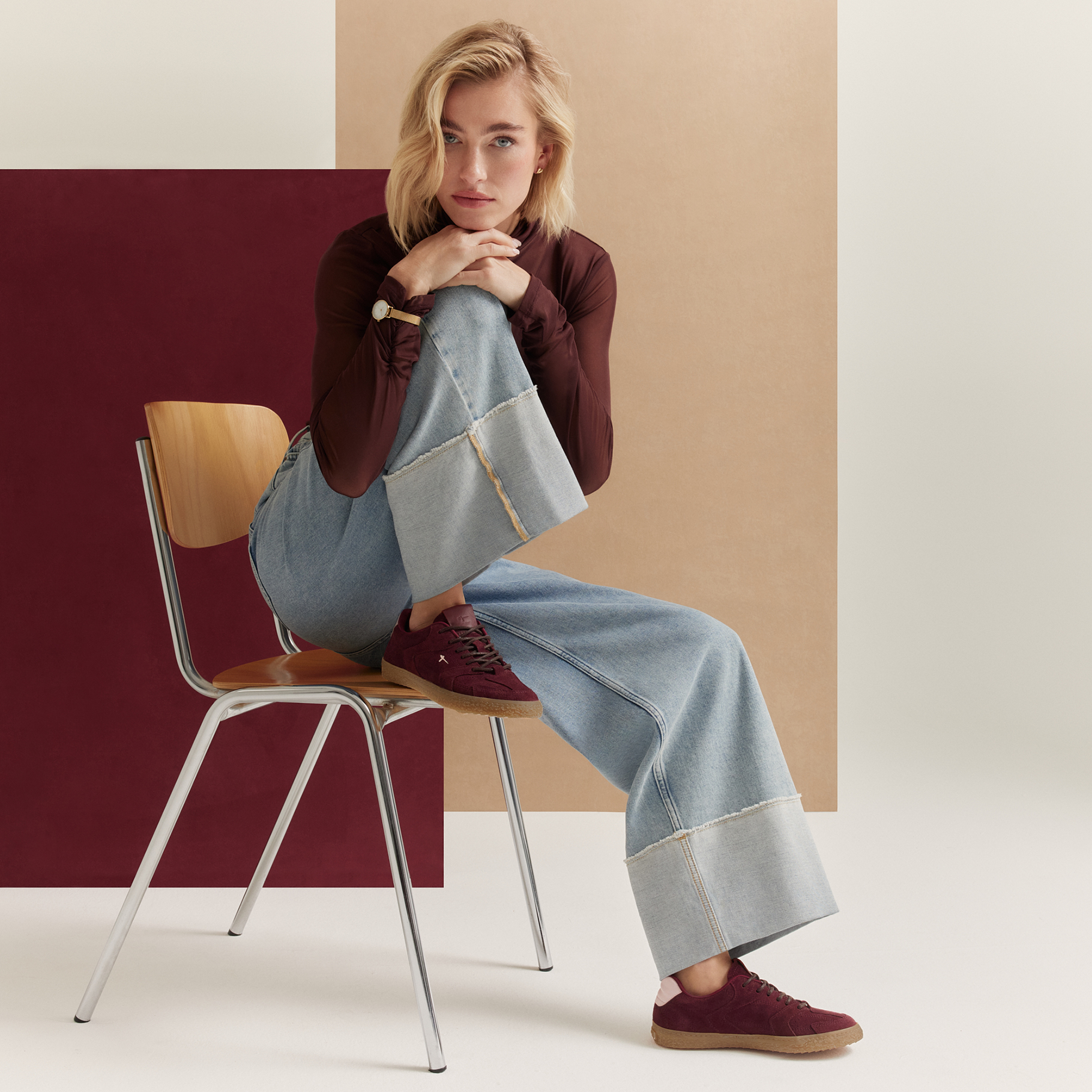 Woman sitting on a wooden chair wearing wide jeans, a burgundy top and sneakers, in front of a geometric background in burgundy and beige.