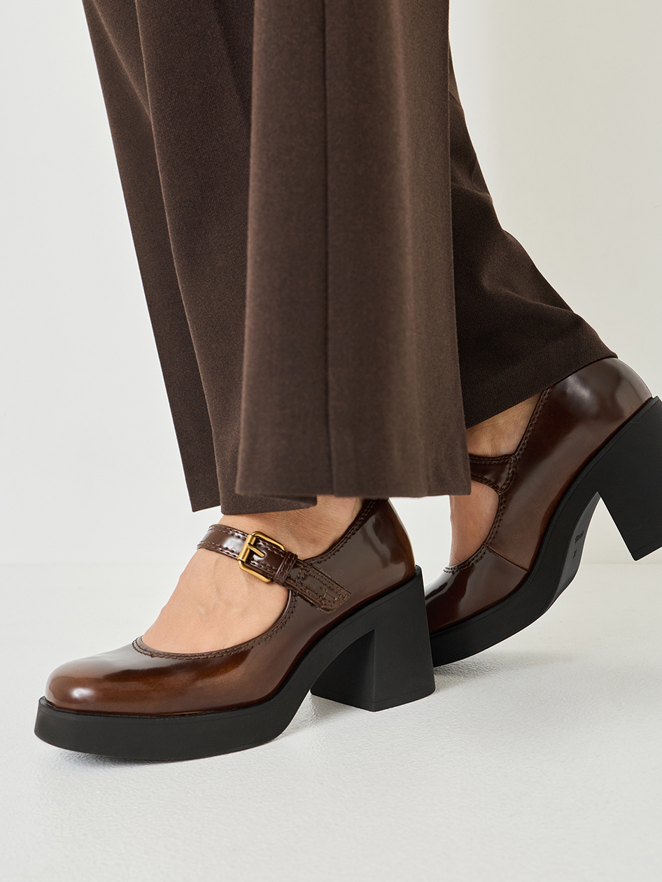 Braune Pumps mit Plateausohle und Schnalle, getragen zu dunklen Hosen, auf weißem Hintergrund fotografiert.