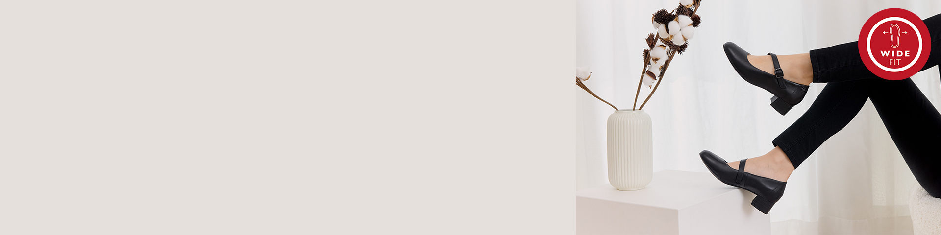 Schwarze Mary-Jane-Schuhe mit breitem Schnitt, getragen zu schwarzen Str&uuml;mpfen, neben wei&szlig;er Vase mit getrockneten Blumen und rotem Wide-Fit Logo oben rechts