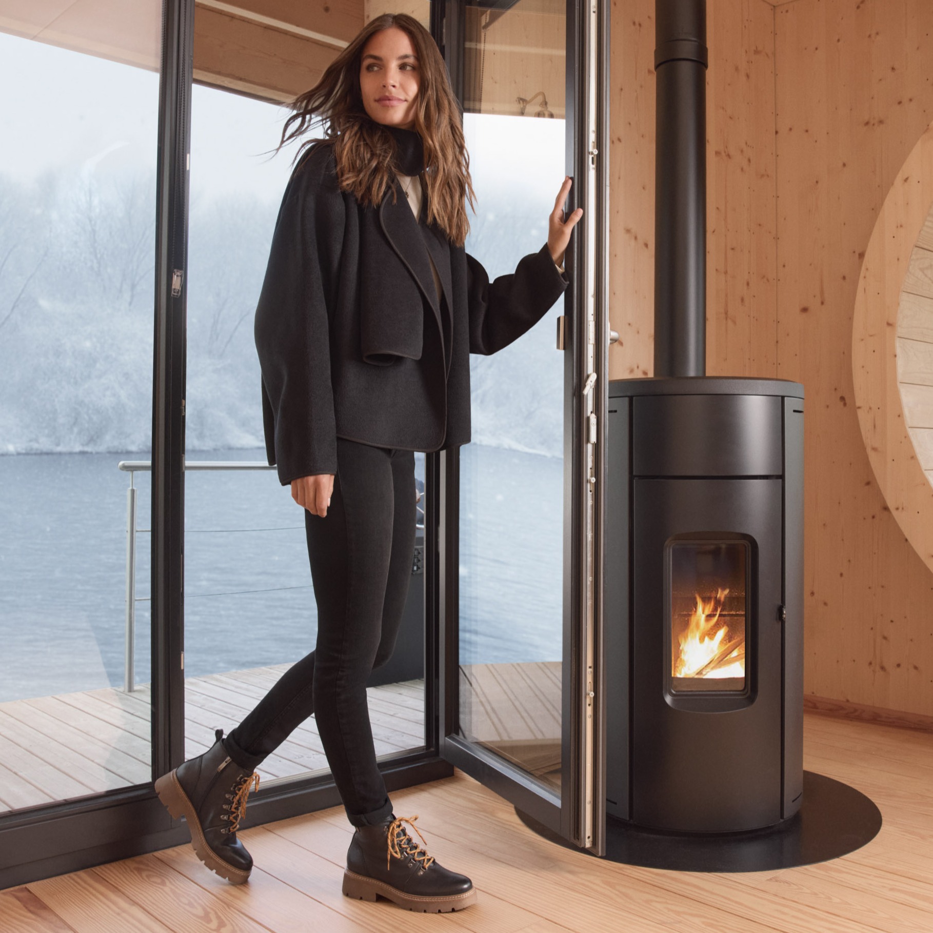 Vrouw in een zwarte outfit staat naast een moderne houtkachel in een lichte houten kamer met panoramische ramen en uitzicht op de bergen