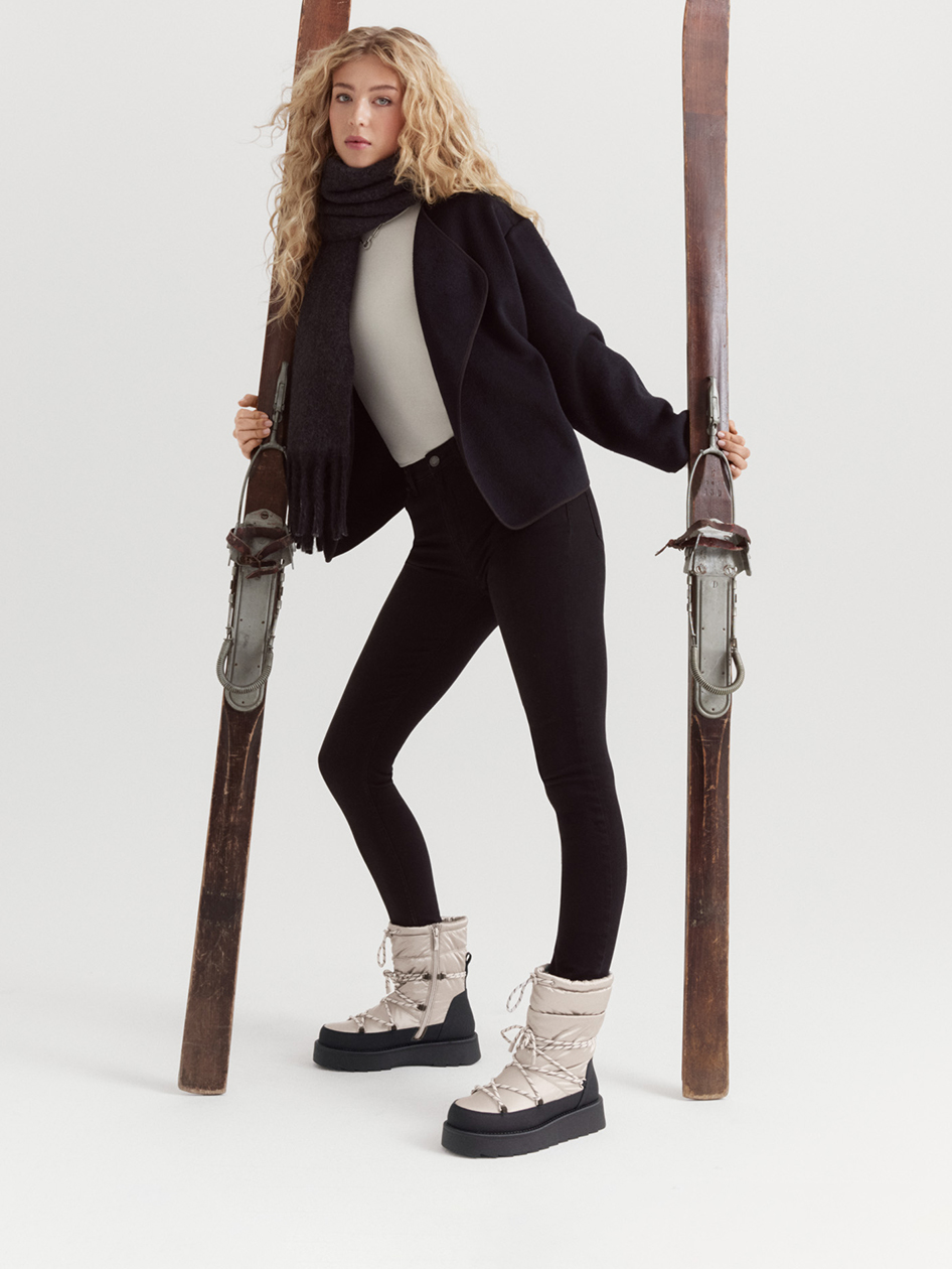 Modèle en tenue d'hiver avec veste noire, leggings et bottes de neige blanches posant entre des skis en bois vintage sur fond blanc.