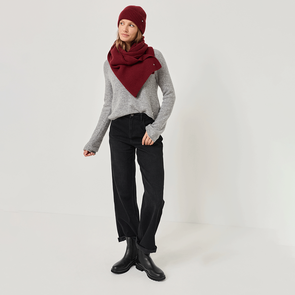 Winteroutfit met een grijze trui, zwarte jeans, bordeauxrode muts en sjaal en zwarte Chelsea boots tegen een lichte achtergrond.