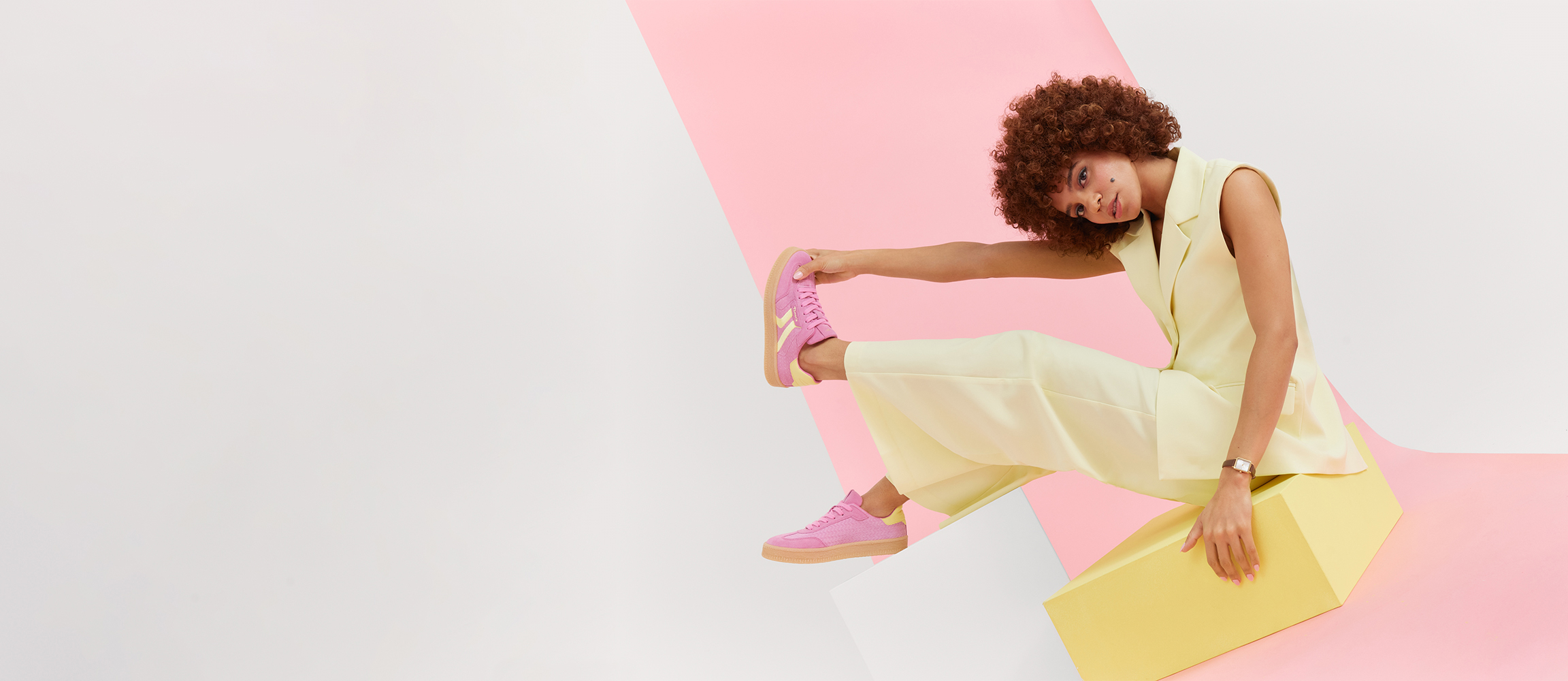 Woman with curly hair in white top and jeans makes a playful pose against pink background, wears pink sneakers.