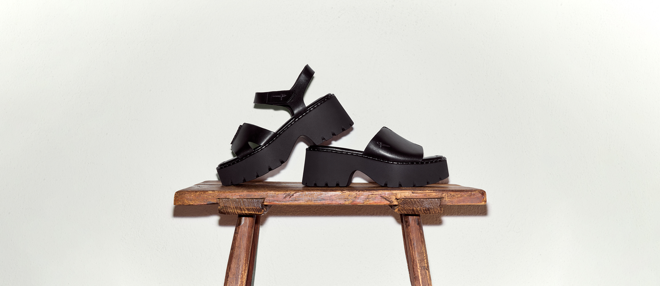 Black platform sandals with a chunky treaded sole on a rustic wooden stool against a light background.