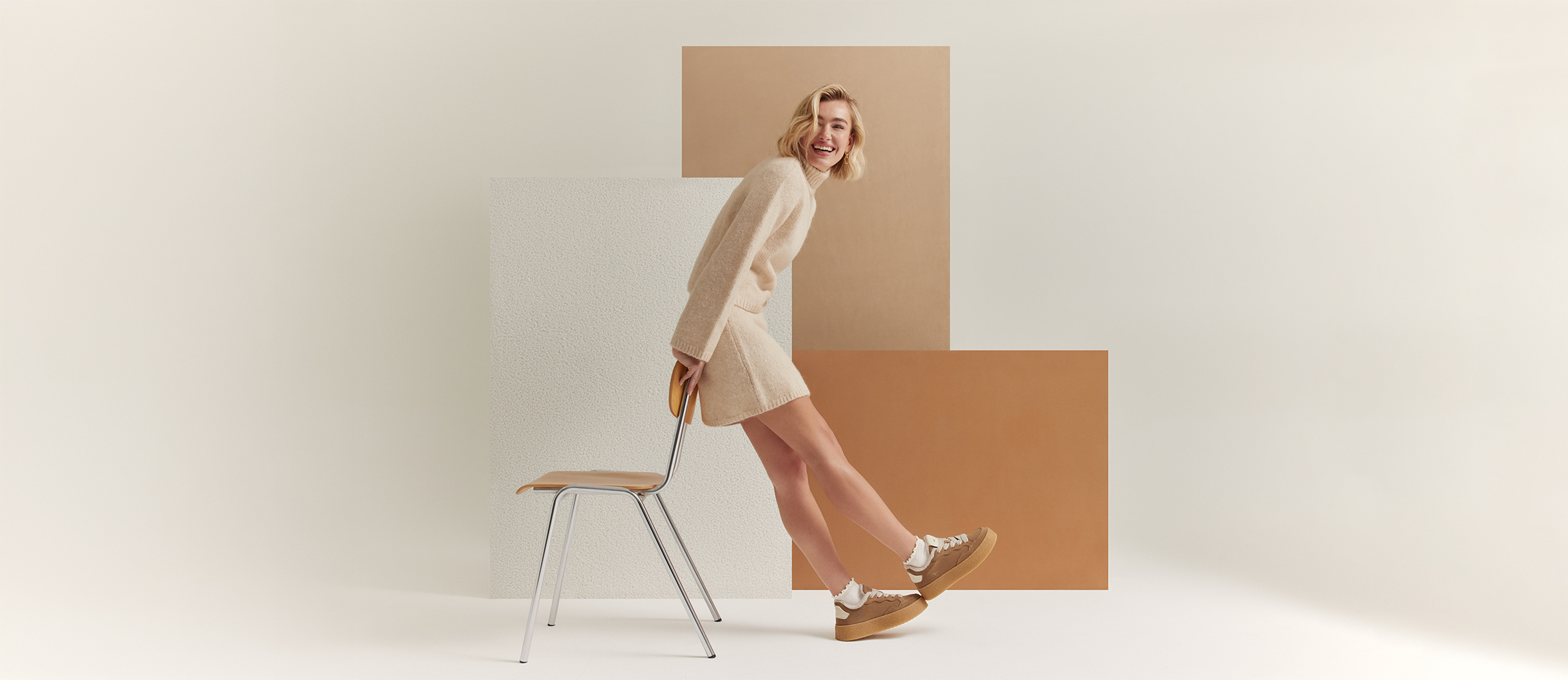 Smiling woman in a beige knitted outfit and brown trainers leaning against a chair in front of a geometric backdrop in earthy tones.