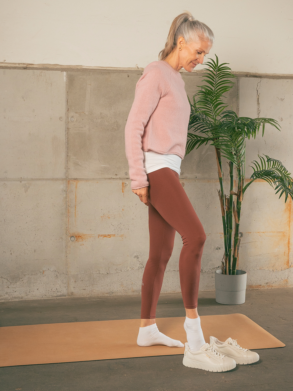Woman in pink sweater and brown leggings stands on yoga mat next to houseplant, wears white sneakers