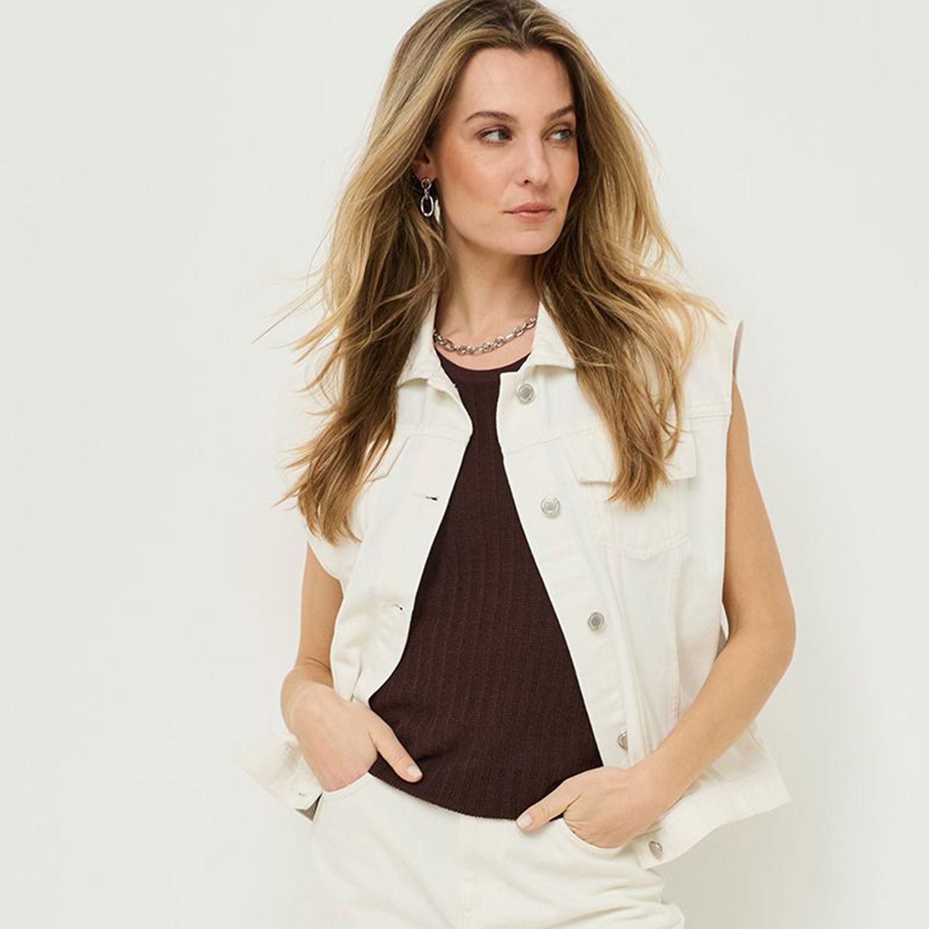 Elegante ritratto di una donna con lunghi capelli biondi, con gilet in denim bianco e top marrone scuro, e collana in argento, su sfondo bianco.