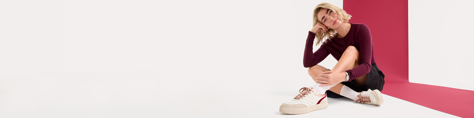 Mod&egrave;le en pull-over bordeaux et short noir assis sur un sol blanc sur fond rose, portant des baskets couleur cr&egrave;me.