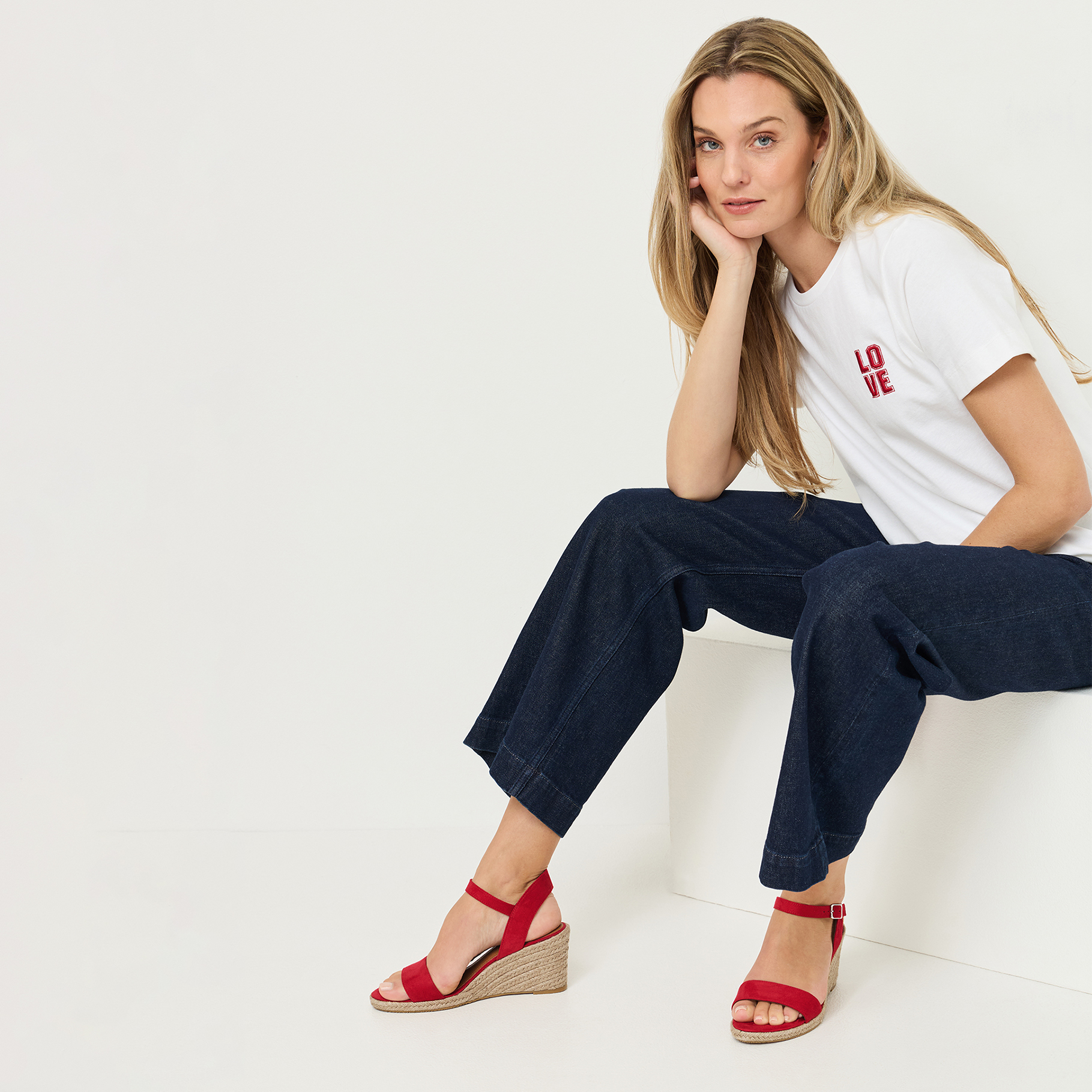 Frau sitzt in wei&szlig;em LOVE-T-Shirt, dunkler Jeans und roten Keilsandaletten auf einem wei&szlig;en W&uuml;rfel.