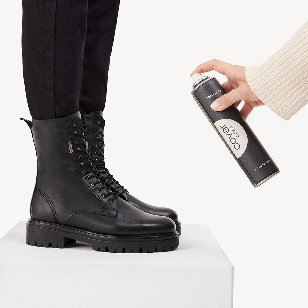 Hand sprüht Schuhpflegespray auf schwarzen Schnürstiefel aus Leder, der auf weißem Podest steht.