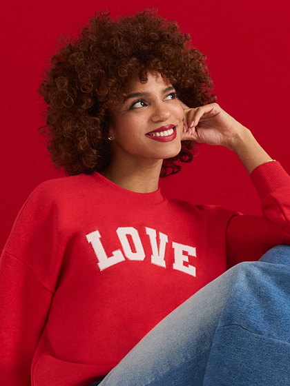 Smiling person with curly hair wears a red LOVE sweater against a red background.