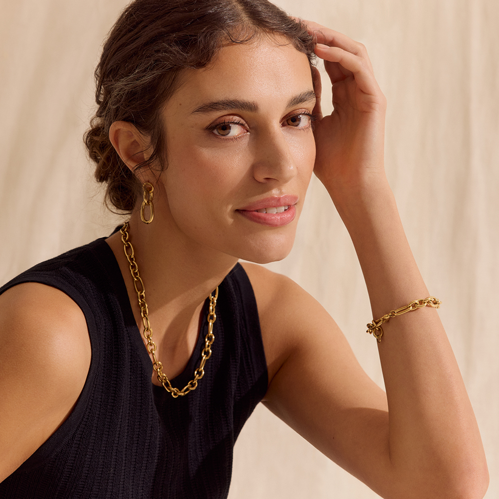 Frau mit goldener Halskette, Armband und Ohrringen, schwarzem Top, l&auml;chelnd vor beigem Hintergrund.