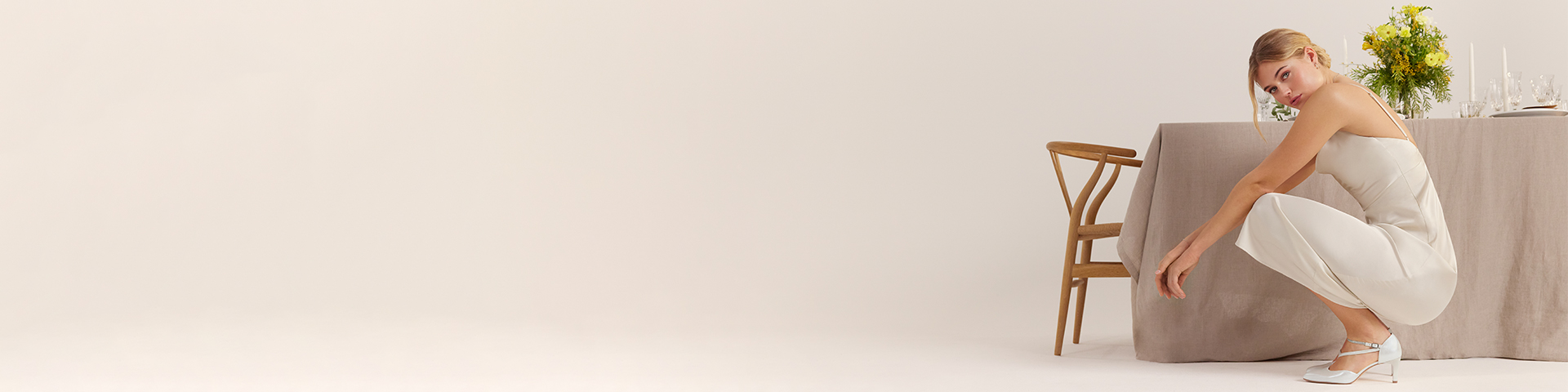 Elegante bruid in een eenvoudige witte jurk hurkt naast een grijze fauteuil, gele bloemen en een houten stoel op de achtergrond.