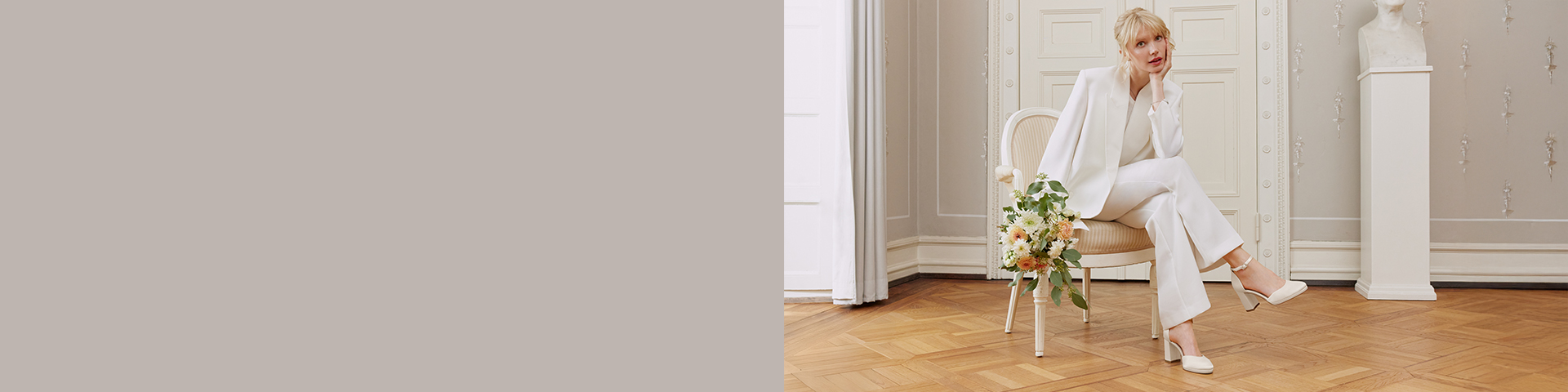 Une mari&eacute;e en &eacute;l&eacute;gant tailleur blanc, tenant son bouquet de mariage, est assise sur une chaise claire dans une pi&egrave;ce minimaliste avec parquet.