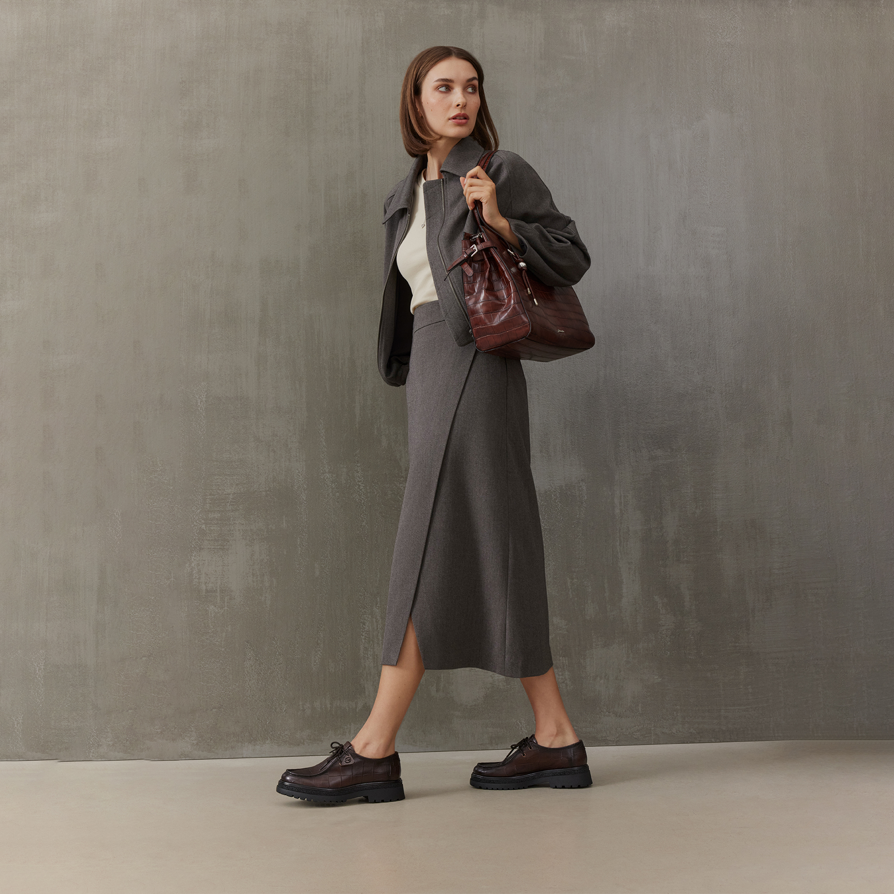 Donna con un outfit grigio composto da gonna midi, giacca e top bianco, indossa una borsa in pelle bordeaux e scarpe stringate scure.
