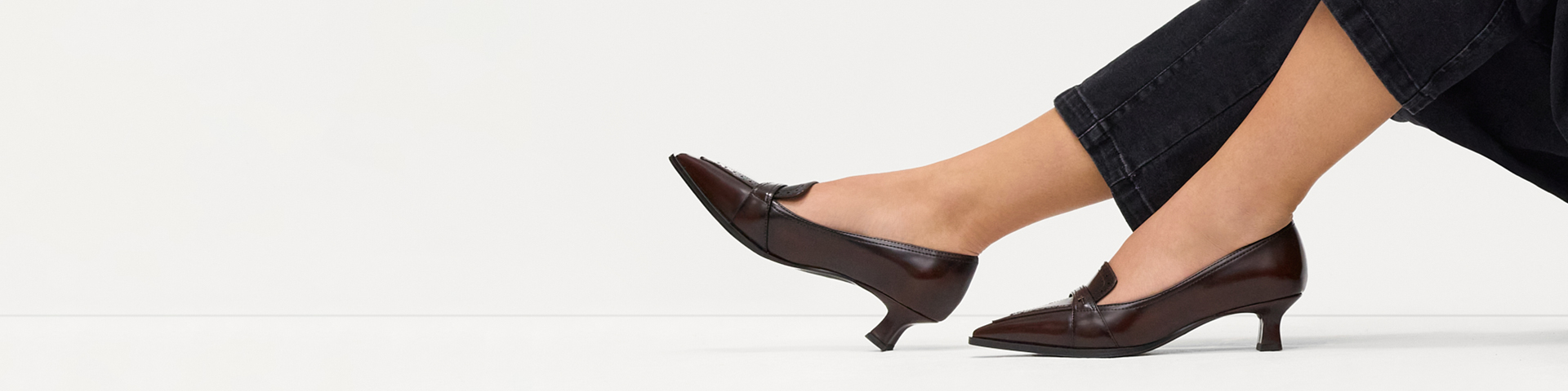 Woman&rsquo;s legs in black jeans wearing dark brown kitten‑heel pumps with a pointed toe against a white background.