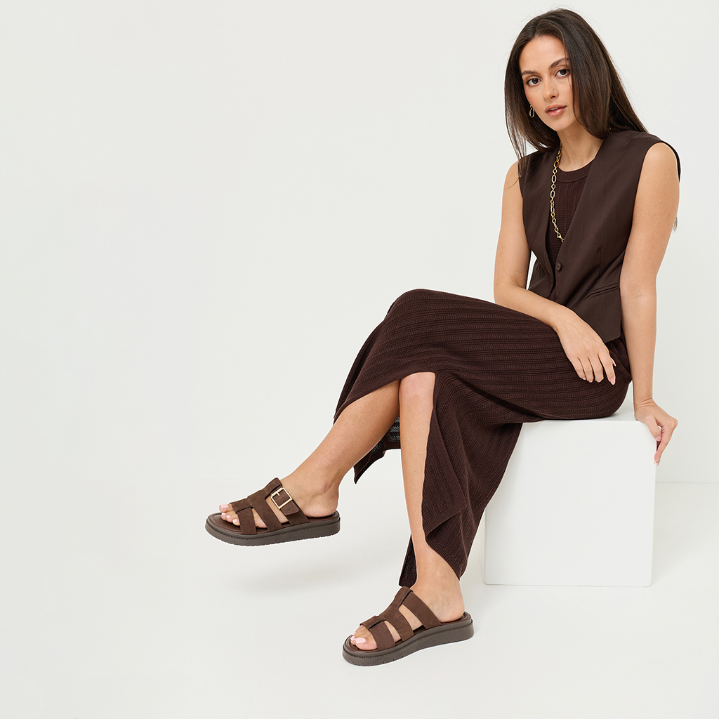 Frau in braunem Kleid und Weste sitzt auf wei&szlig;em W&uuml;rfel, tr&auml;gt braune Sandalen mit Schnalle und goldene Halskette.