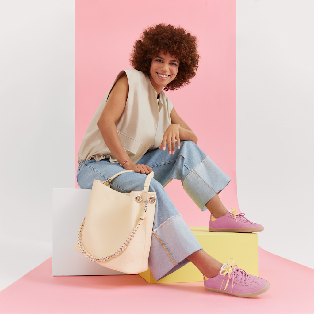 Moderne outfit med beige bluse, jeans, rosa sneakers og hvid taske foran en pastelfarvet baggrund.