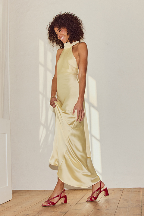 Woman in a cream-colored silk dress and red sandals dances smiling in front of a white wall.