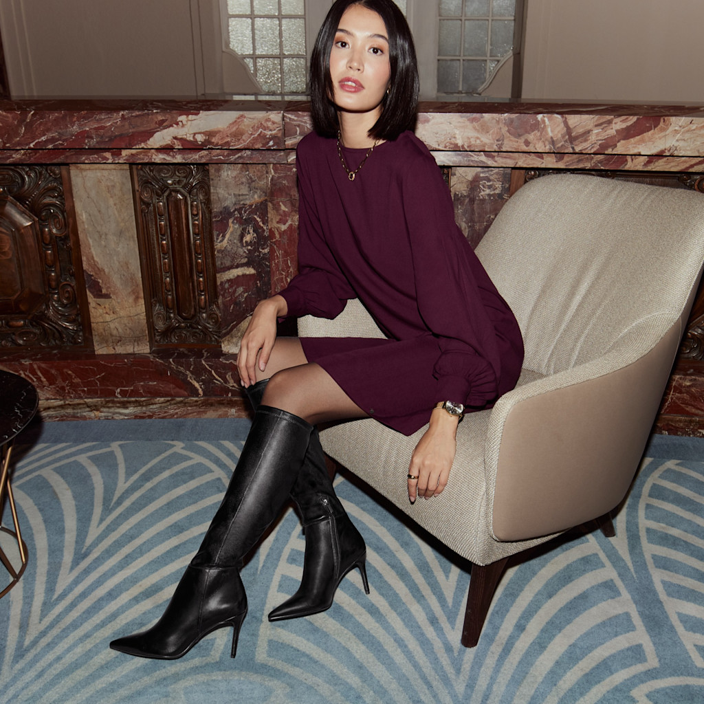 Portrait de mode élégant: femme en robe violet foncé et bottes noires assise sur un fauteuil beige devant un mur de marbre et un tapis à motifs.