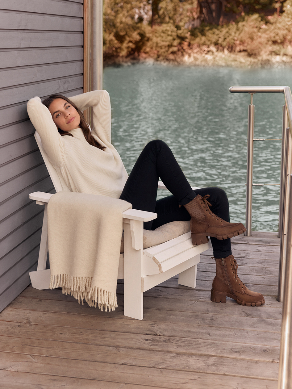 A woman in a cream sweater and brown boots sits relaxed on a white chair by the lake with a blanket.