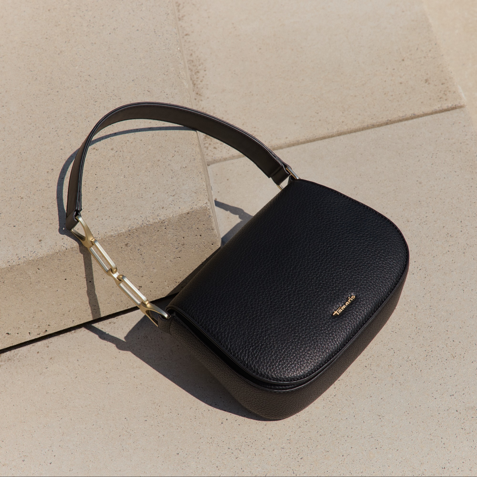 Elegant black leather handbag with golden logo and metal details on light concrete background in sunlight.
