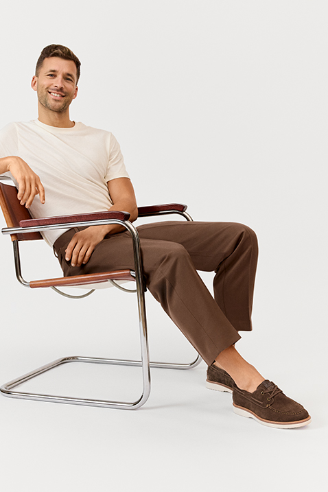 A person wearing a white T-shirt and brown trousers sits relaxed in a vintage cantilever chair with wooden armrests.