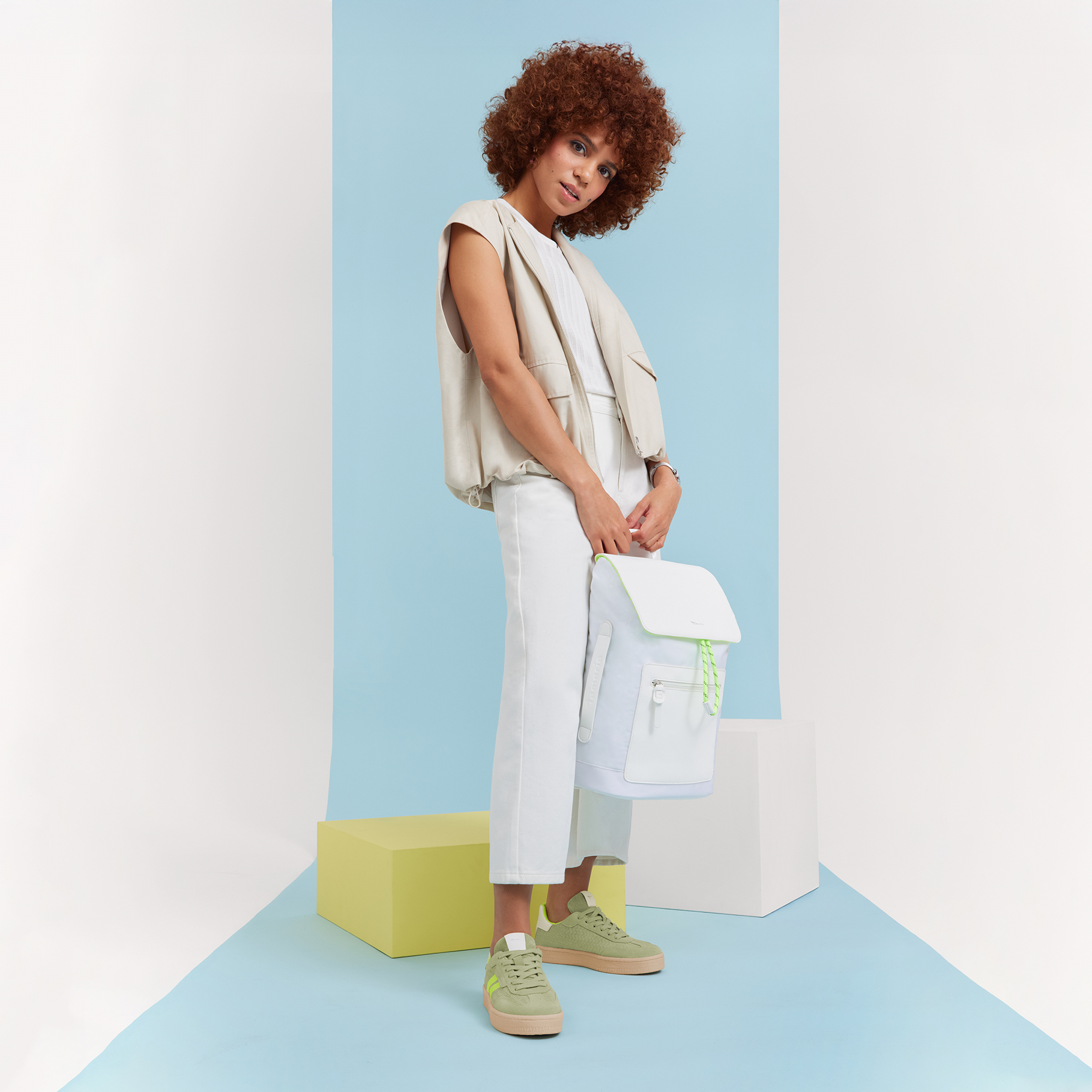 Model in beige top en witte broek poseert voor lichtblauwe achtergrond, draagt mintgroene sneakers en heeft krullend bruin haar.