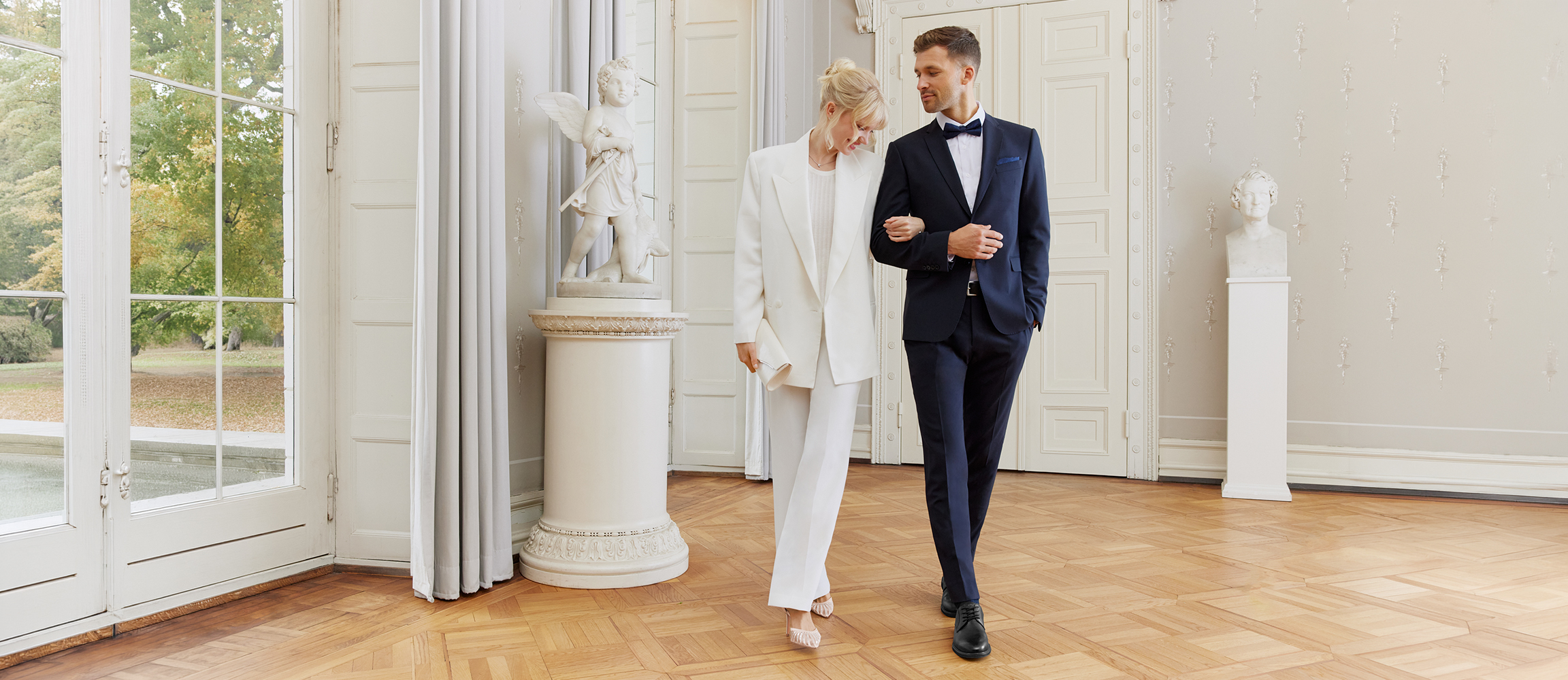 Elegantn&iacute; svatebn&iacute; p&aacute;r v b&iacute;l&eacute; a tmavě modr&eacute; stoj&iacute; v světl&eacute;, klasick&eacute; m&iacute;stnosti s vysok&yacute;mi okny a sloupy.