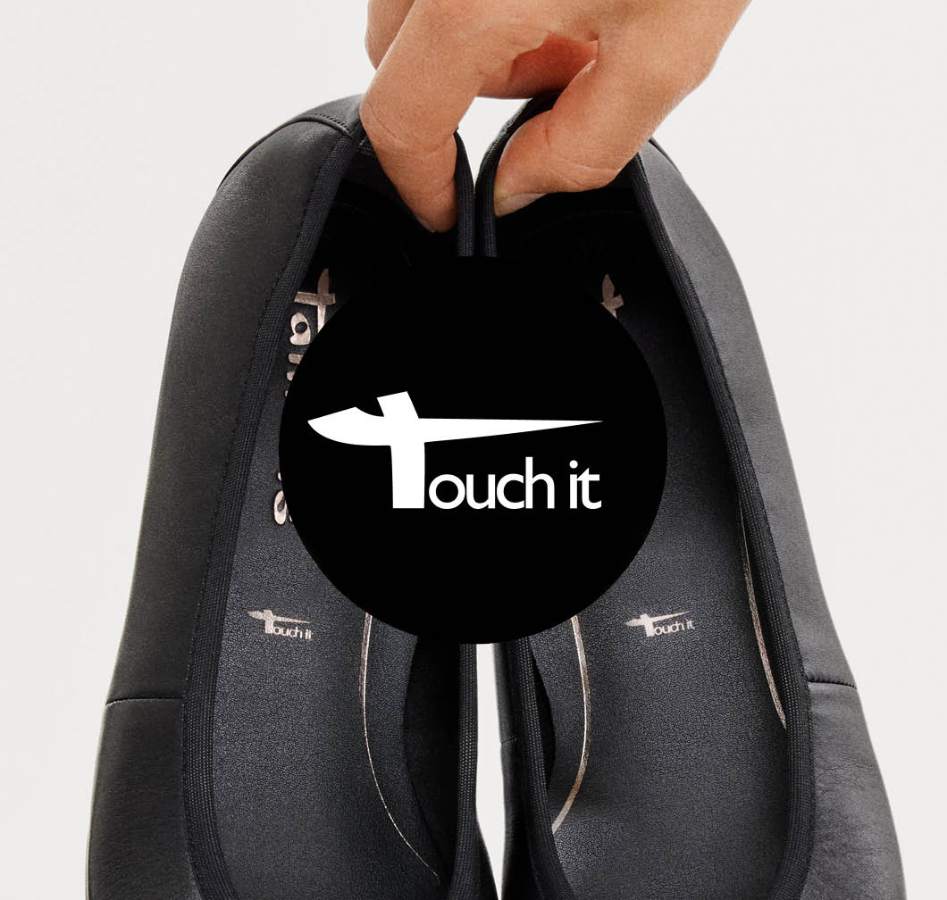 A hand holding a pair of black shoes with the insole visible, with a black circle featuring the Touch-it logo in the foreground.