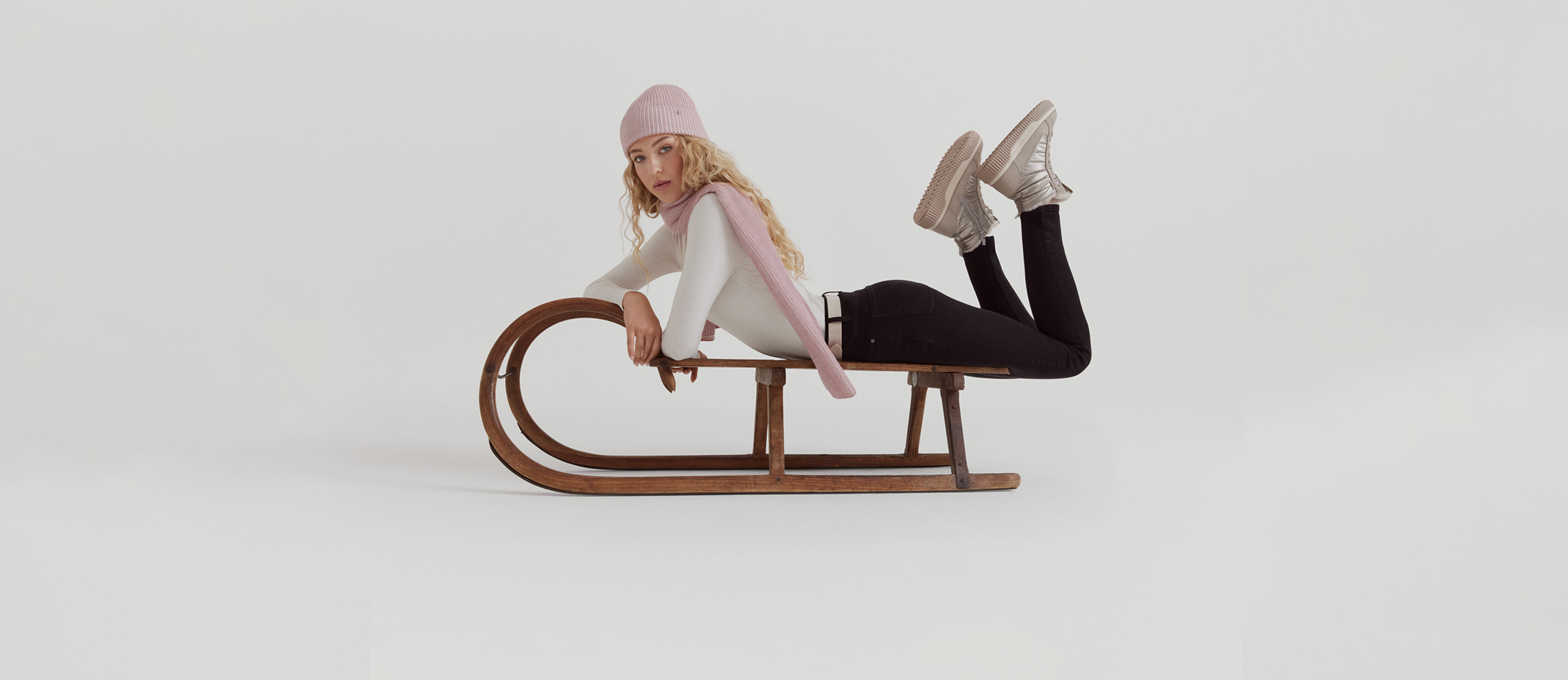 Modell in Winterkleidung posiert auf einem vintage Holzschlitten vor weißem Hintergrund, trägt rosa Mütze, Schal und weiße Stiefeletten