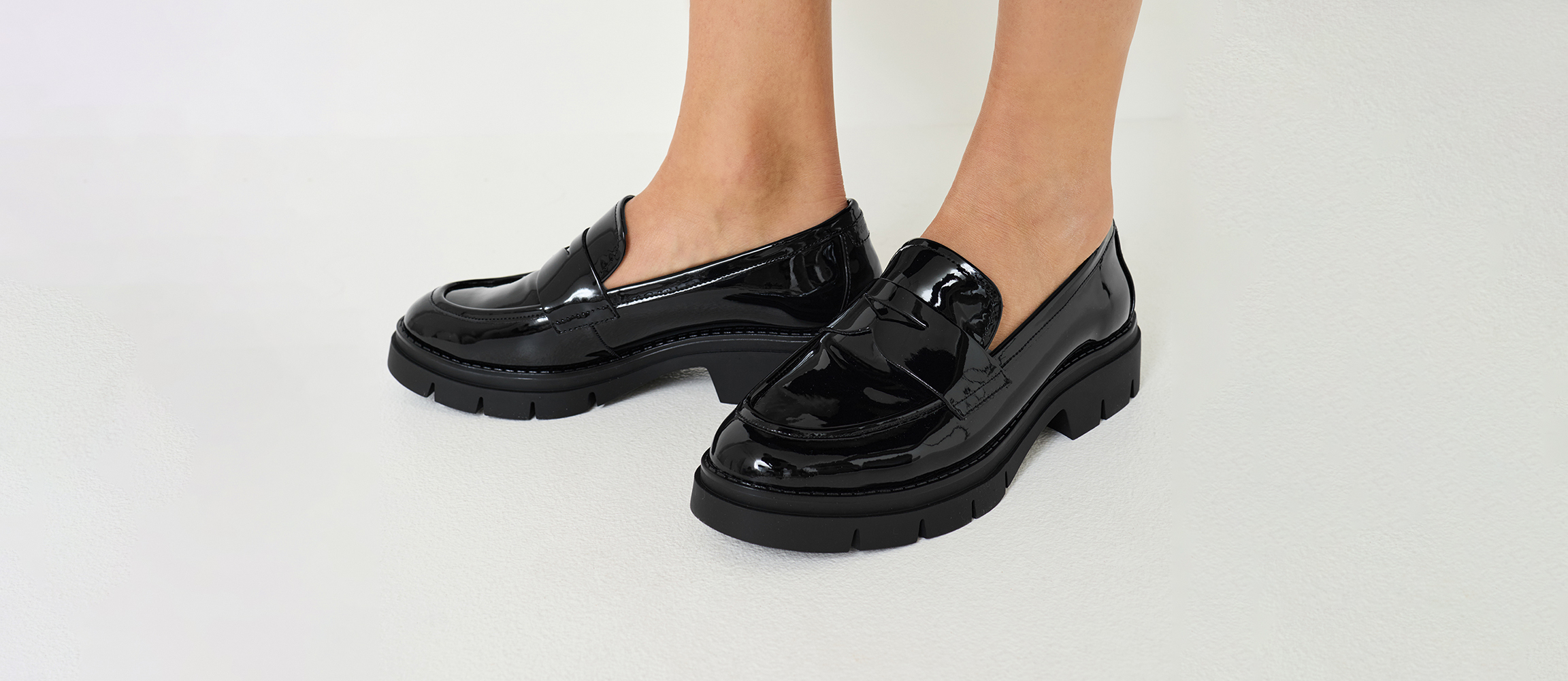 Zwarte lakloafers met een robuuste plateauzool, gedragen aan blote voeten voor een witte achtergrond.