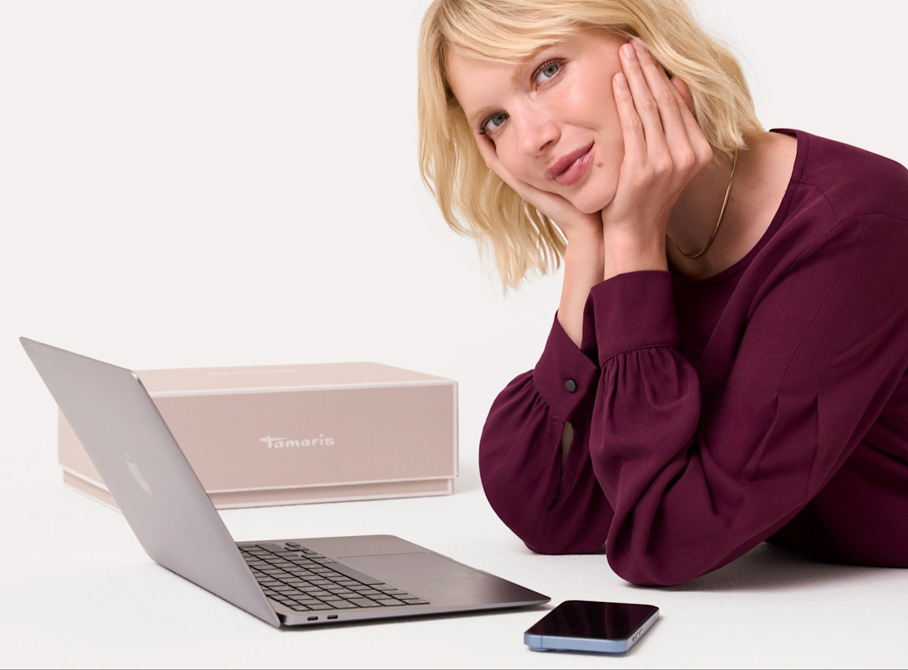 Nachdenkliche Person in burgunderroter Bluse vor Laptop und Tamaris-Schuhkarton, Hand an Wange gelehnt, auf wei&szlig;em Hintergrund.