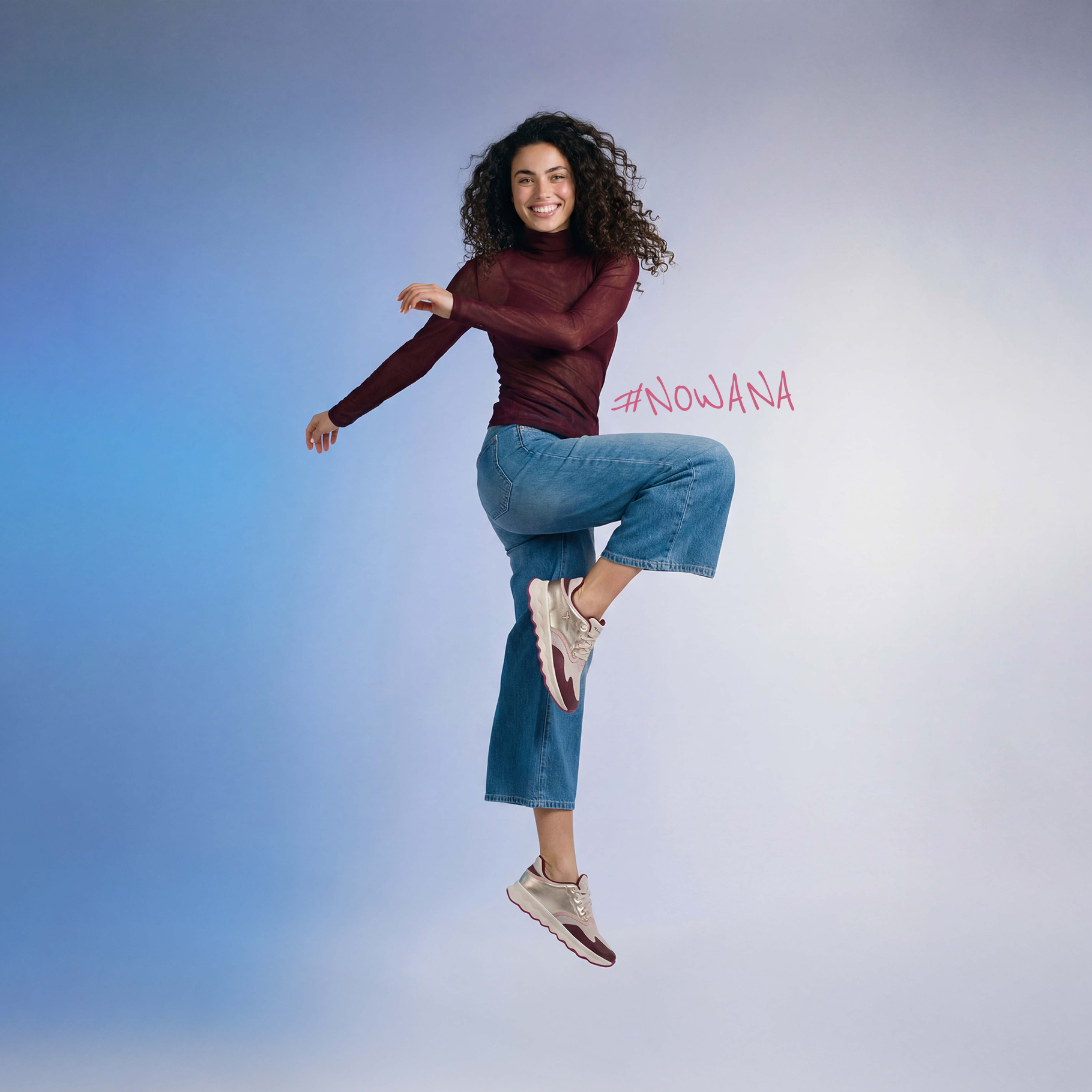 Donna sorridente salta in aria, indossa una maglietta bordeaux, jeans blu e scarpe da ginnastica beige. Hashtag #NowANA sullo sfondo.
