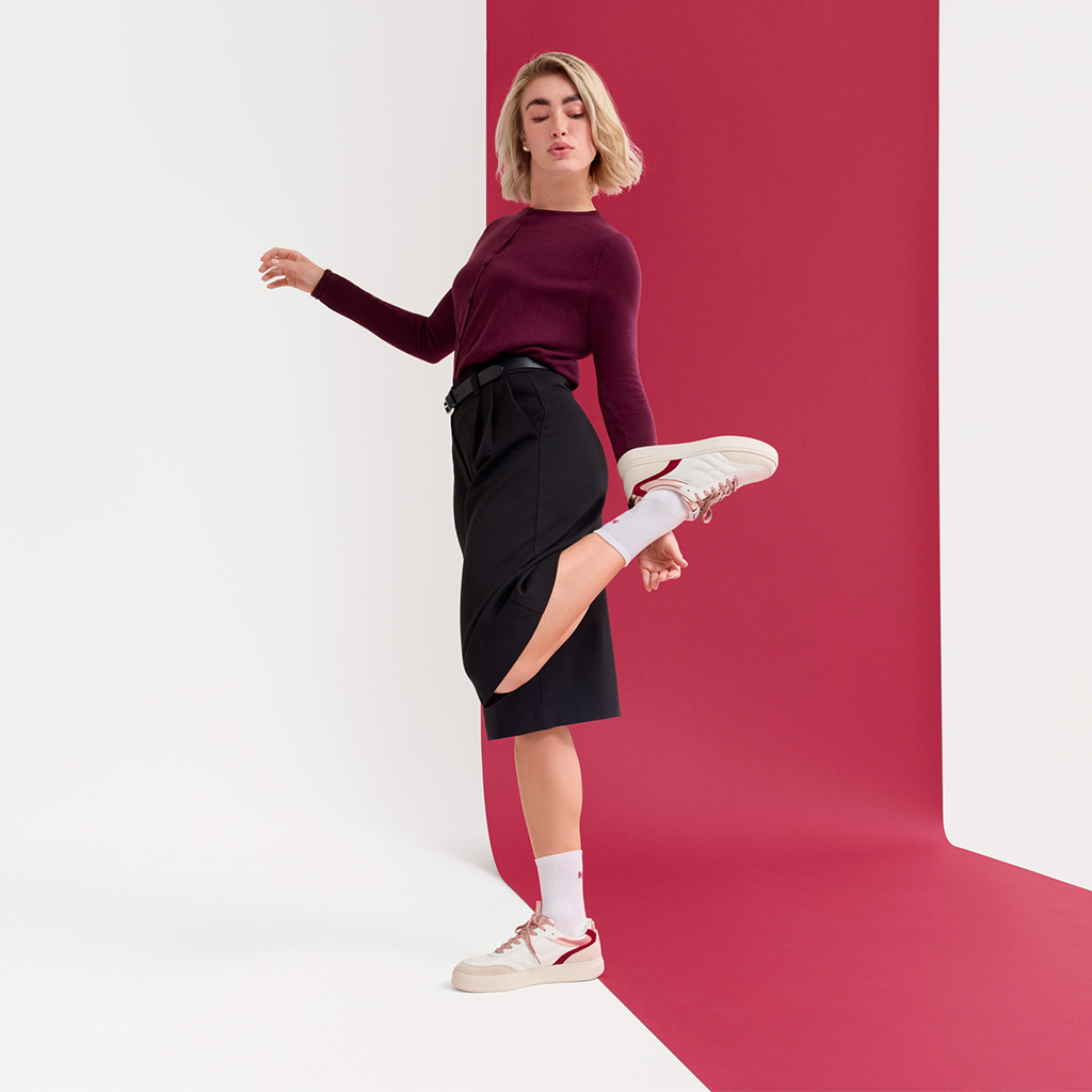 Une femme blonde vêtue d'un pull-over bordeaux et d'une jupe noire pose de manière enjouée sur un fond rose et porte des baskets blanches.