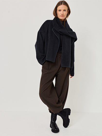 Une personne vêtue d'un manteau court noir et d'un pantalon large marron pose sur fond blanc dans des bottines noires.