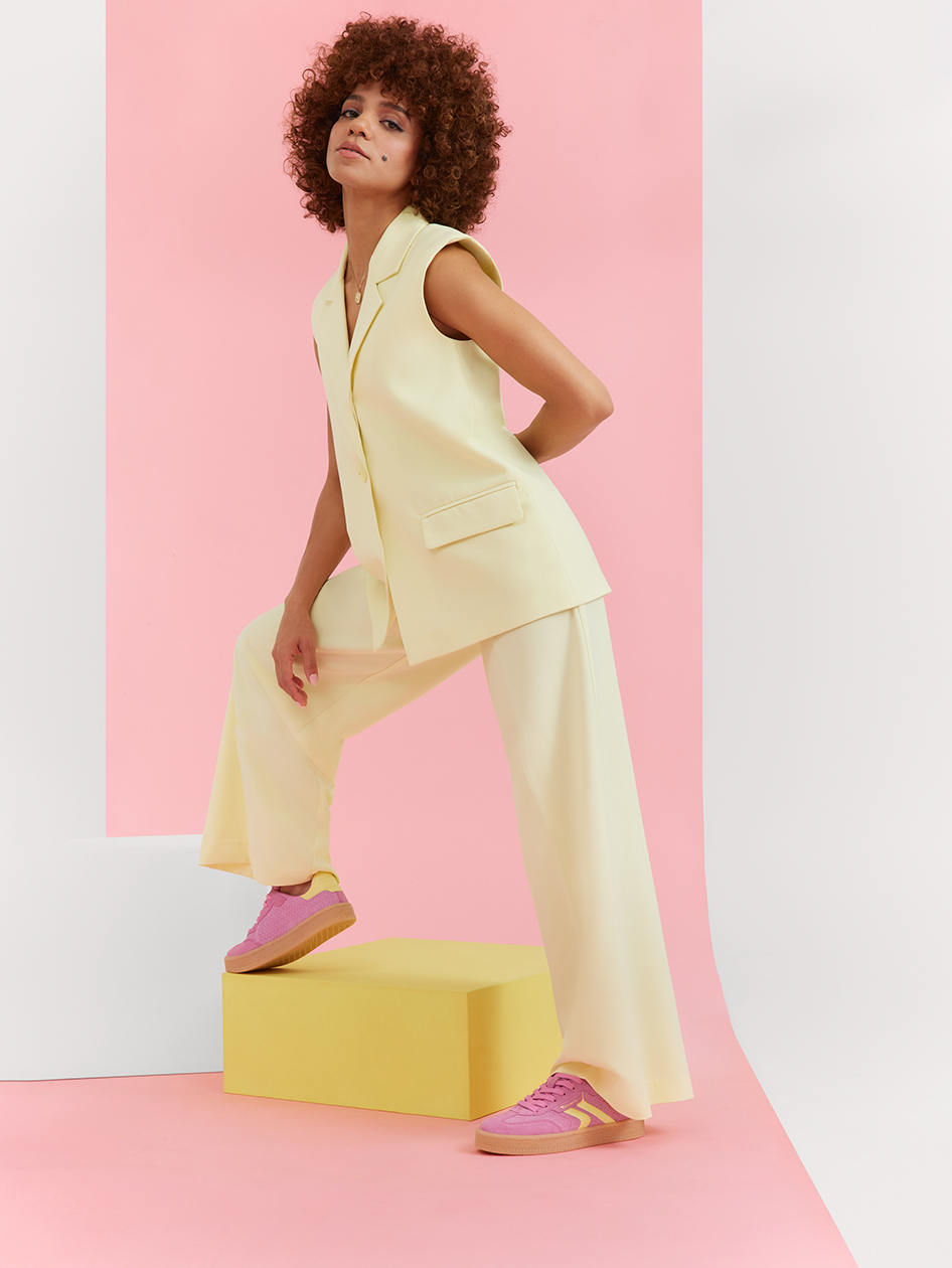 Model in a cream-colored pantsuit and pink sneakers poses on a yellow pedestal against a pink background, with curly hair.