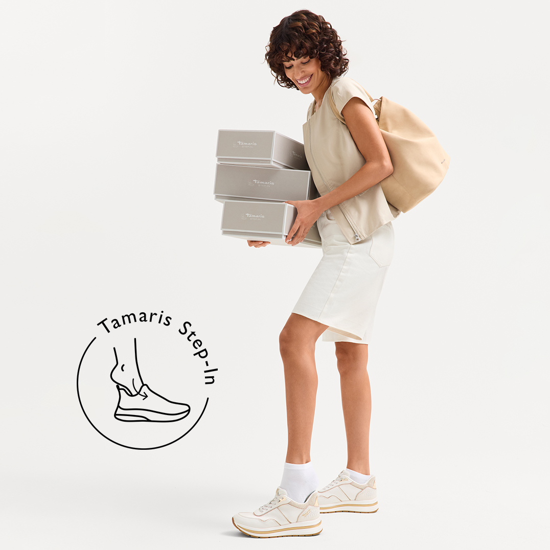 Personne en tenue claire portant plusieurs bo&icirc;tes &agrave; chaussures Tamaris en souriant, tout en enfilant des baskets blanches.