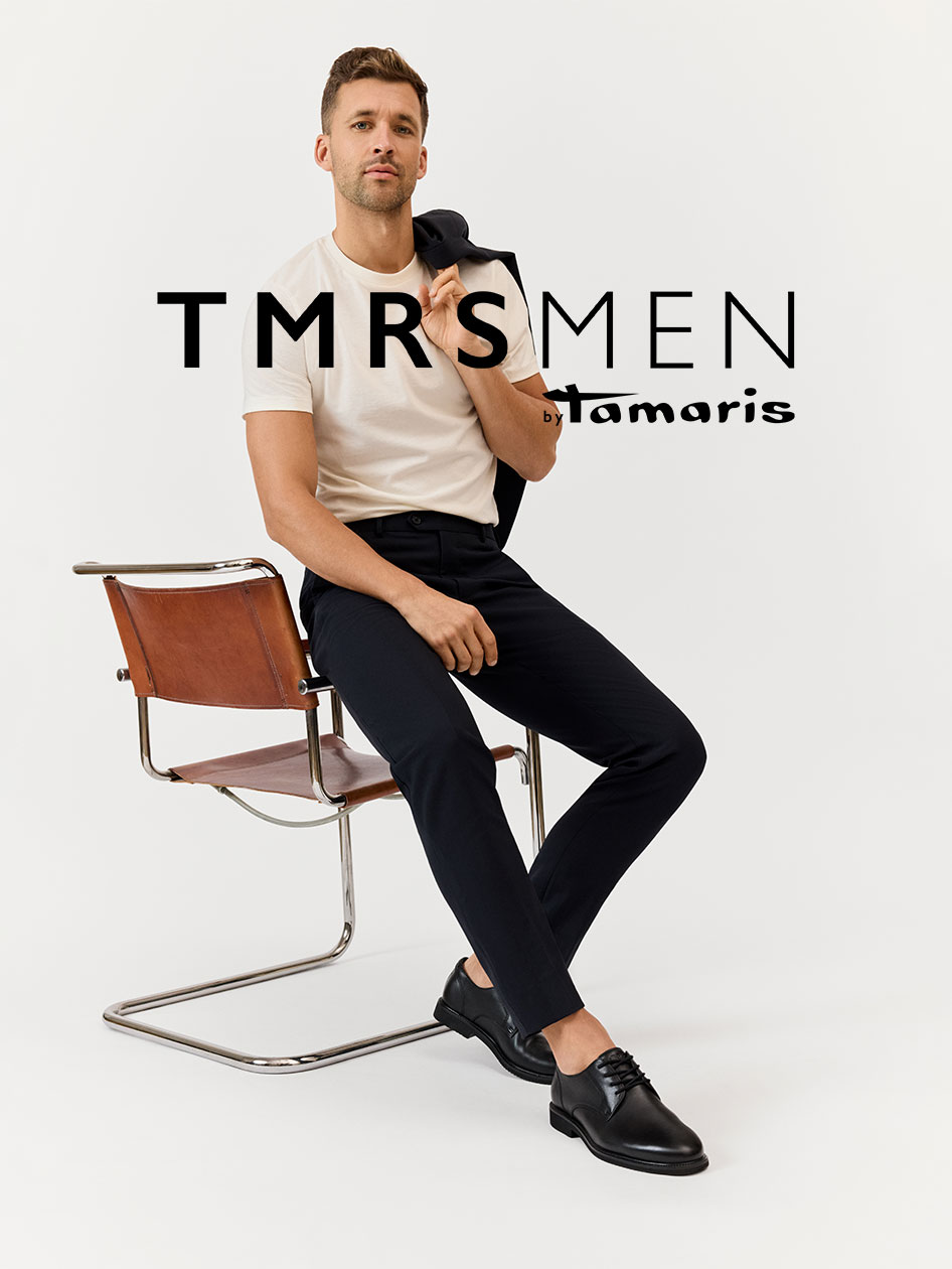 Modello uomo in T-shirt bianca e pantaloni neri seduto su una sedia in pelle marrone, indossa scarpe in pelle nere. Logo TMRS MEN visibile.