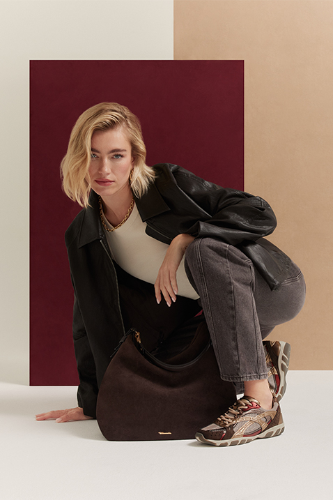 Frau in schwarzer Jacke, grauer Jeans und bunten Sneakers hockt vor weinrotem Hintergrund.