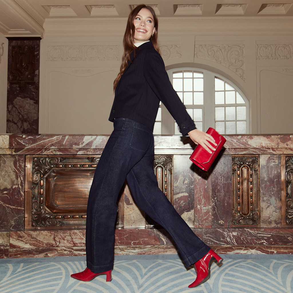 Donna in abito scuro con stivali rossi e borsa posa in modo dinamico in una stanza elegante con pareti di marmo.