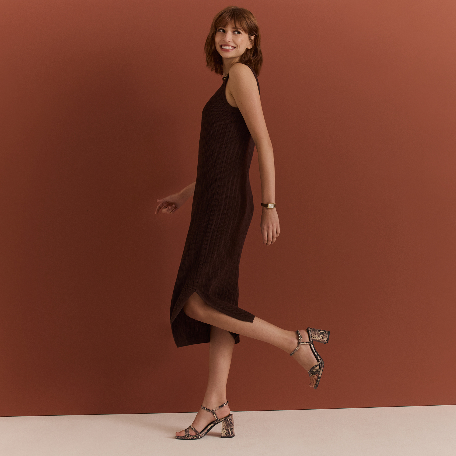 Eine Frau in einem eleganten schwarzen Kleid und Schlangenleder-Sandalen posiert l&auml;chelnd vor einem rostbraunen Hintergrund.