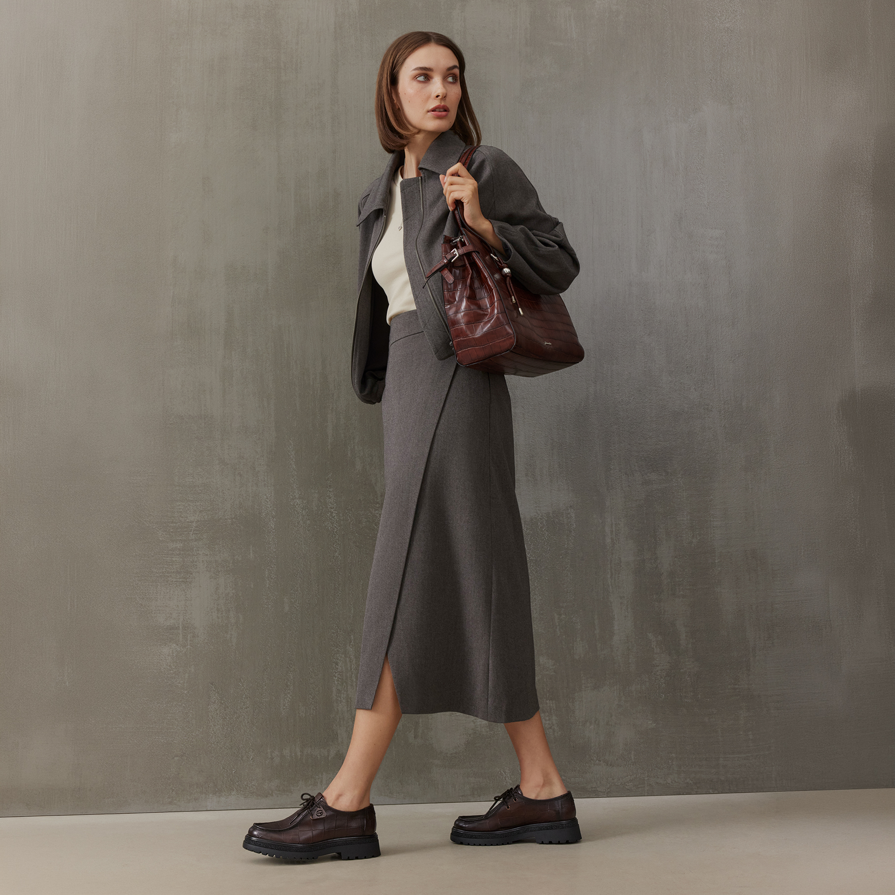 Vrouw in een bruine jas en midi‑rok draagt een donkerrode handtas en bruine veterschoenen tegen een grijze achtergrond.