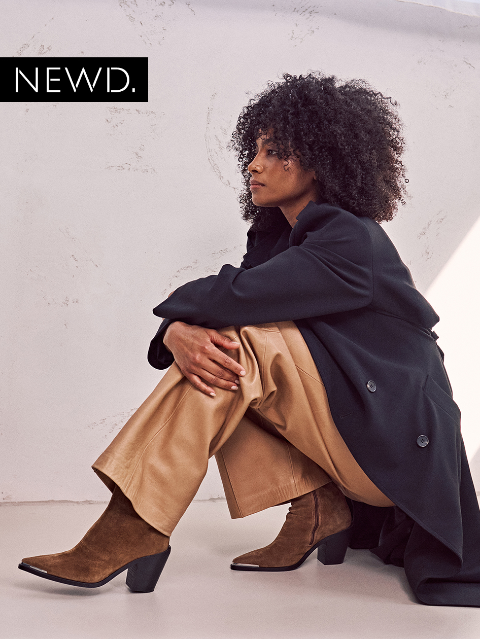 The model in a black coat, beige trousers and brown boots sits on the side against a white background, with curly hair in profile.