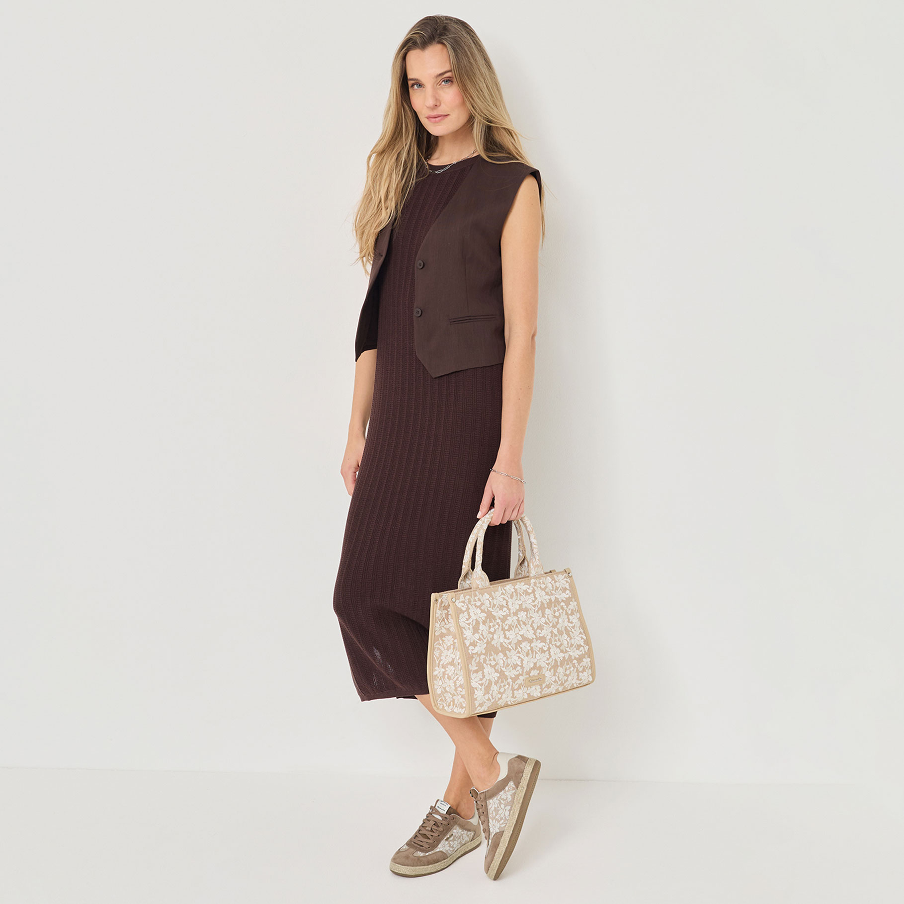 Elegant brun kl&auml;nning med v&auml;st, kombinerad med beige handv&auml;ska med blomm&ouml;nster och matchande sneakers mot vit bakgrund.
